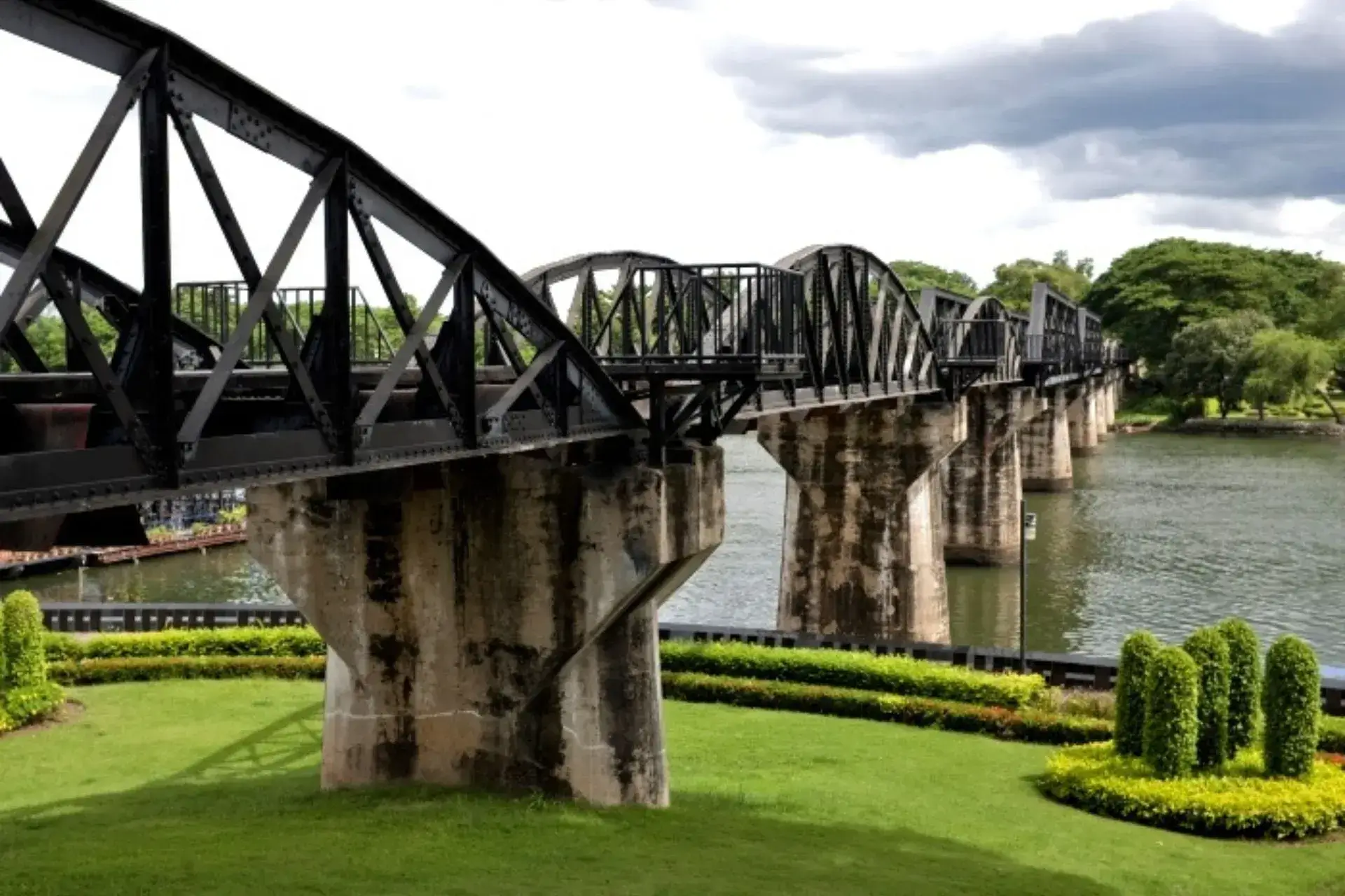 Parte del puente sobre el río Kwai de hierro en la parte superior y cemento en la inferior. Hierba en primer plano y vegetación al fondo