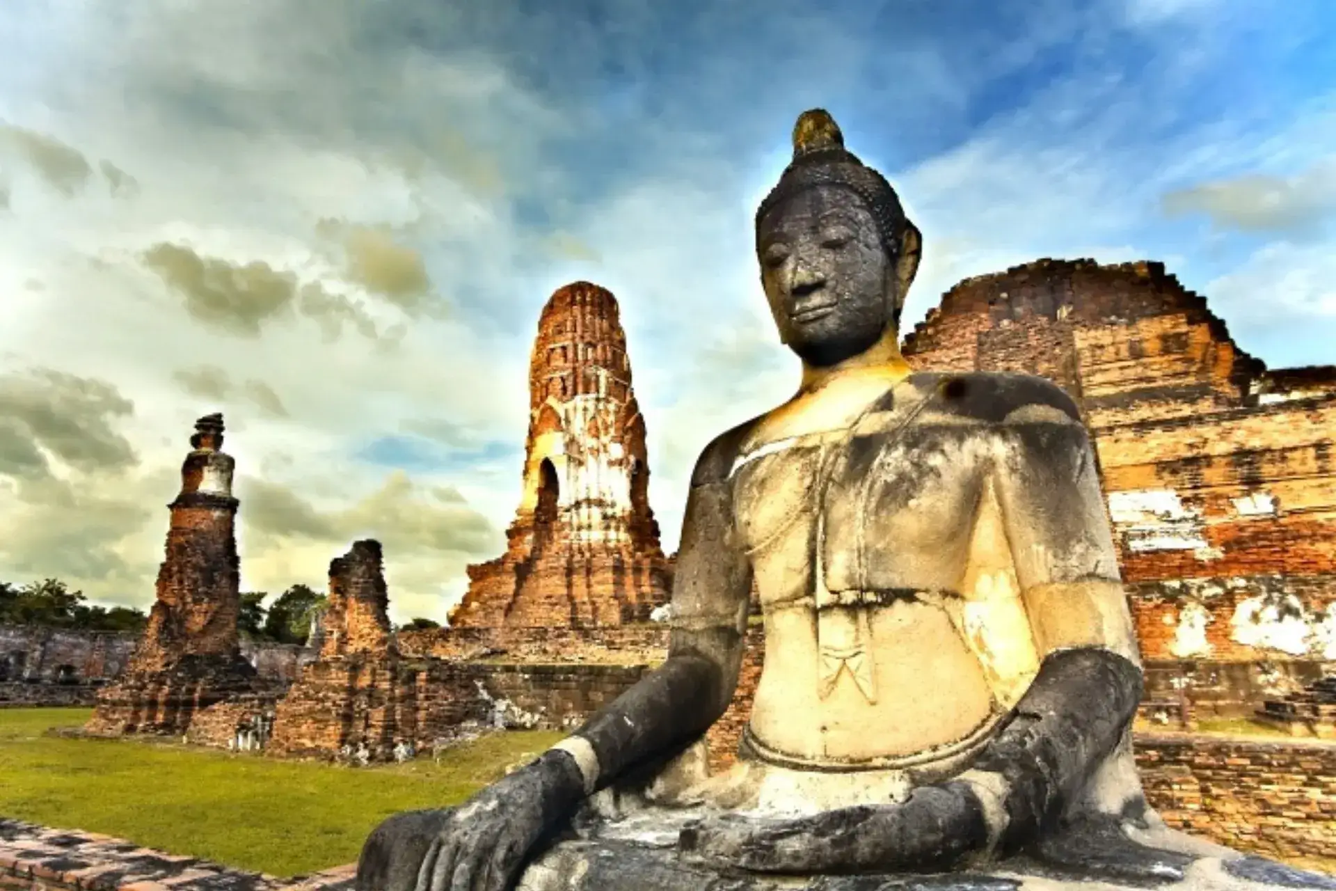 Escultura de Buda de gran tamaño y restos de antiguas construcciones a su espalda bajo un cielo azul y grisáceo