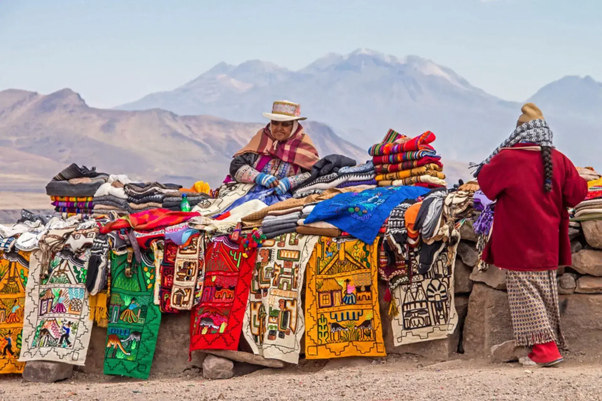 Dos mujeres indígenas venden tejidos tradicionales en un puesto con las montañas al fondo