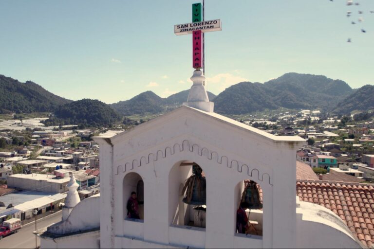 Foto del campanario de una iglesia y de fondo casas del pueblo y las montañas