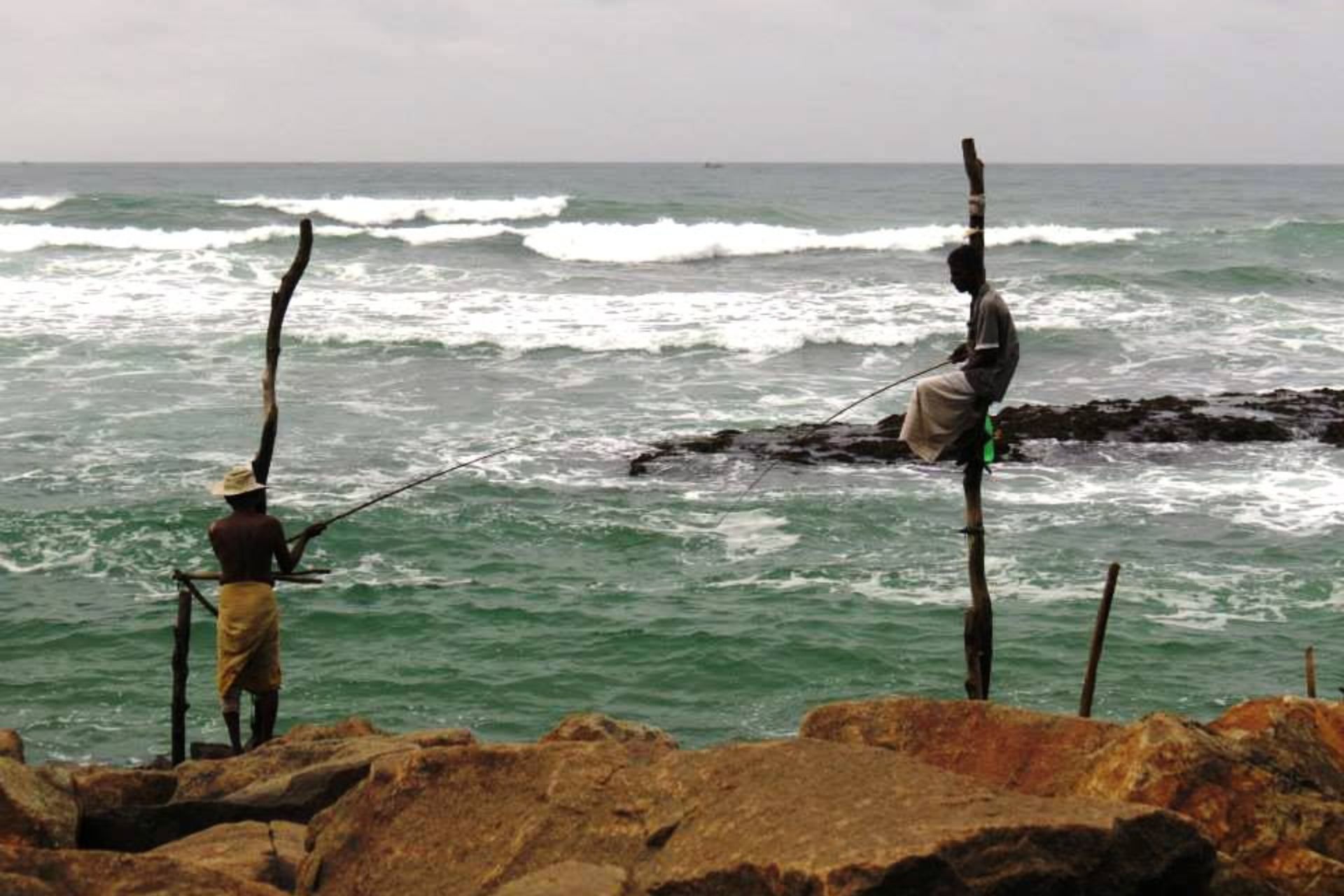 Mar y rocas, un hombre en lo alto de un palo sosteniendo caña de pescar. Otro palo y al lado otro hombre con su caña de pescar